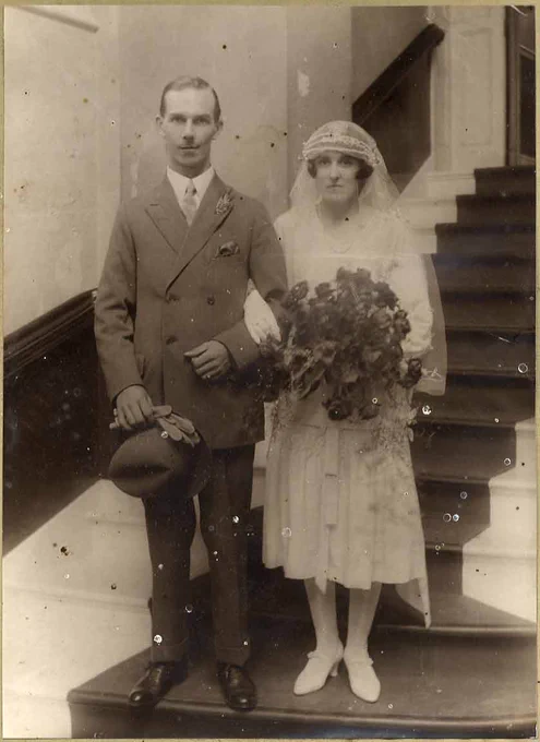 Black and white photograph of a newly married couple, c. 1930. They stand at the foot of a staircase. The groom wears a double-breasted suit with a sprig of white heather in the button-hole, and carries a pair of gloves and a trilby hat. The bride is dressed all in white, with a veil, and holds a bouquet of roses.