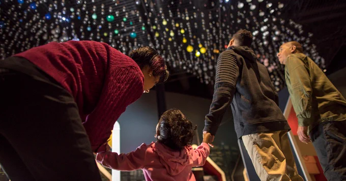 Une famille se tenant par la main et regardant vers le plafond rempli de boules lumineuses suspendues dans un espace d'exposition sombre.