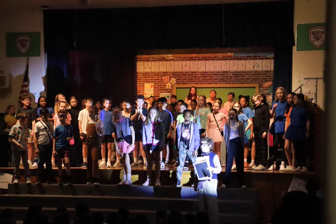 A large group of children performing on a school stage with a brick wall backdrop and spotlight on a boy holding a book.