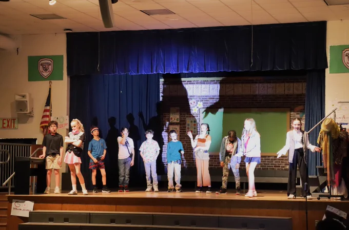 Children performing a school play on stage with a backdrop of a brick wall and blue curtains under stage lights.