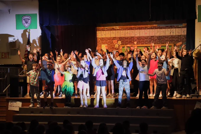 A diverse group of children enthusiastically performing on stage with raised arms in a school auditorium.