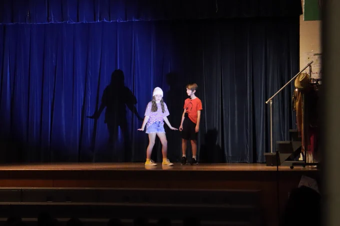Two children perform on a dimly lit stage with a dark blue curtain backdrop, casting large shadows behind them.