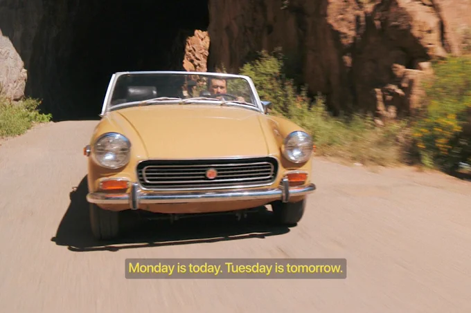From Pluribus, Carlos-Manuel Vesga as Manousos drives a yellow convertible surrounded by rocks and bushes. Text on the bottom middle of the screen reads “Monday is today. Tuesday is tomorrow.”