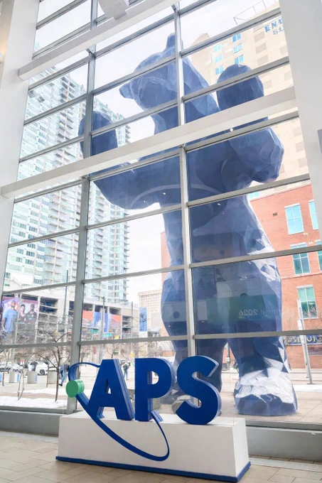 A giant blue bear sculpture peers through the glass windows of the Colorado Convention Center. Inside the lobby, large APS letters sit on a white display stand in front of the windows, with downtown buildings visible outside.