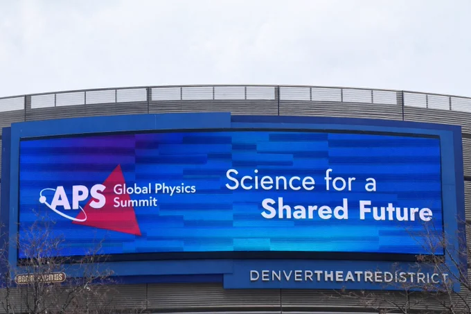 A large curved digital billboard on a building in the Denver Theatre District displays “APS Global Physics Summit – Science for a Shared Future” against a blue background under a cloudy sky.