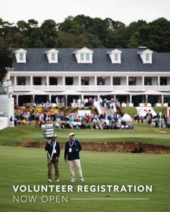 Golf course scene with two individuals standing on the green carrying scoreboard signs. A large clubhouse is in the background with numerous people on the course. Text in the foreground reads "Volunteer Registration Now Open.