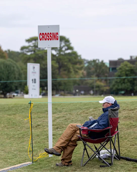 A person sits on a folding chair beside a golf course path, near a sign labeled "CROSSING." They are wearing a white cap and outdoor clothing, and appear to be observing the course. The background features the 18th hole marker and greenery.