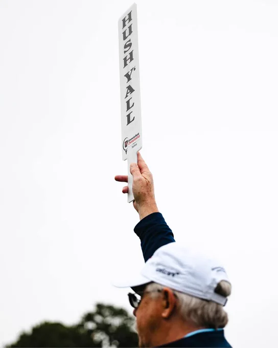 An individual is holding up a paddle that reads "Hush Y’all" against a clear sky. The person is wearing a white cap and dark clothing.