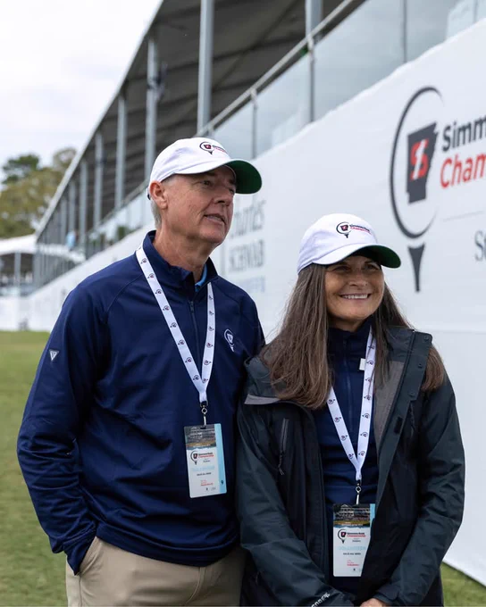 A person wearing a cap and navy attire stands beside another person in similar clothing, both with identification lanyards. They are in front of a white backdrop featuring the Simmons Bank Championship logo.