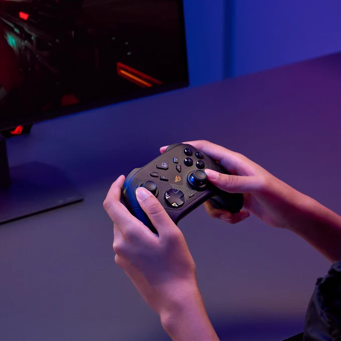 Personne tenant une manette gaming noire ROG Raikiri II Xbox Wireless devant un écran de PC éclairé en rouge, dans un setup gaming aux lumières violettes et bleues.