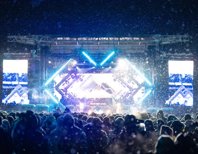 Looking over the heads of a large crowd at an outdoor concert at Ford Park in Vail, Colorado with bright lights around the stage and snow falling.
