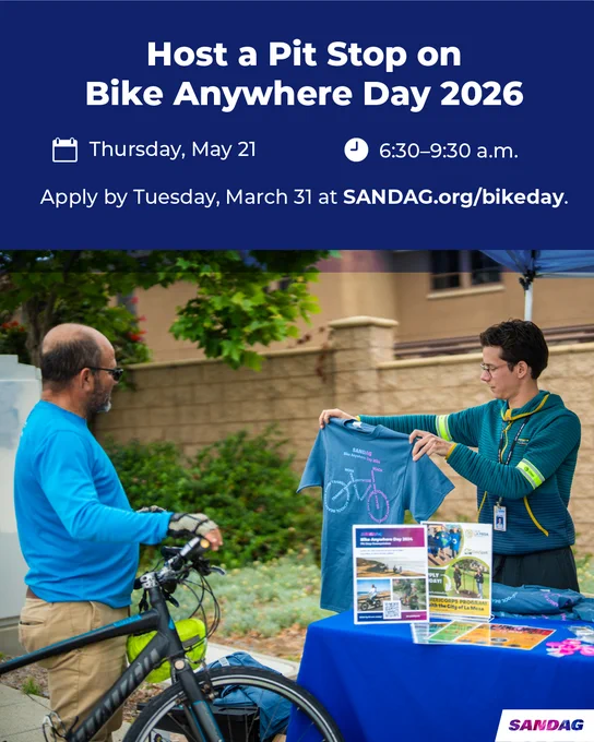 The image shows two people at an outdoor event table, one holding a blue t-shirt with a bicycle graphic. The table displays informational brochures and a sign. Above them, text reads: "Host a Pit Stop on Bike Anywhere Day 2026," with details "Thursday, May 21" and "6:30–9:30 a.m." It mentions applying by March 31 at SANDAG.org/bikeday. The logo "SANDAG" appears at the bottom right.