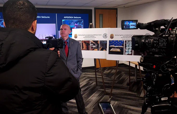 DEA SAC Olesky speaks with Spanish reporters at a press conference. TV camera on right; poster boards in background