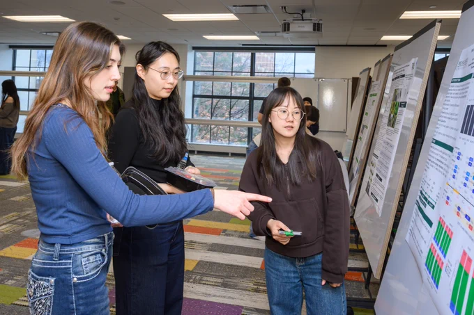 A candid medium shot of two people engaged in a thoughtful discussion in a university lounge or lobby. A woman in a dark blazer and a green "International Studies and Programs" lanyard is smiling and listening intently to a man in a grey suit who is speaking. In the background, other event attendees are visible, along with a large vertical banner for the International Scholar Research Exposition featuring the Michigan State University Spartan helmet logo.