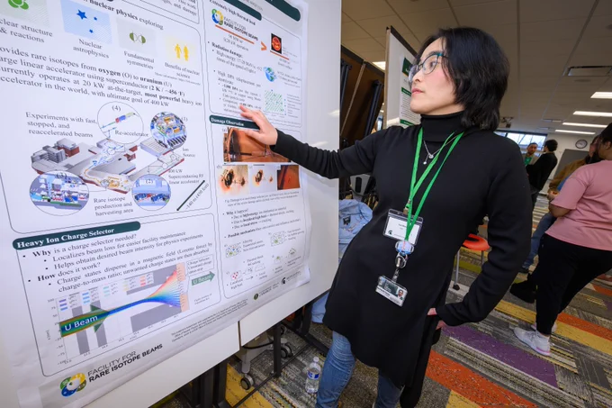 A candid, medium shot of a woman with glasses and a green "International Studies and Programs" lanyard gesturing toward a research poster. The background is a brightly lit, modern university room filled with rows of presentation boards and other attendees, illustrating a vibrant atmosphere of academic feedback and professional networking.