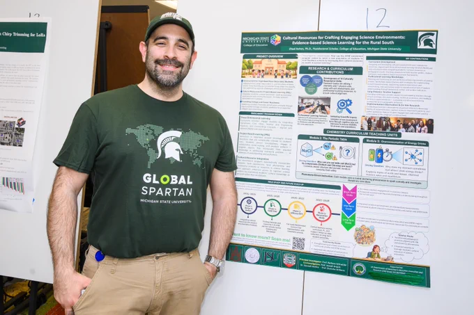 A candid, medium shot of a male scholar wearing a green Global Spartans T-shirt and khaki pants. He is standing beside his research poster.