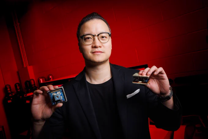Jun Wang, assistant professor of electrical and computer engineering at the University of Nebraska–Lincoln, holds two variants of the novel 10 kilovolt silicon carbide MetaPak switch cells inside his lab in the Scott Engineering Center. He is wearing a dark blue sport coat over a black shirt, with glasses. The lab is lit with red light. (Jordan Opp/University Communication and Marketing)