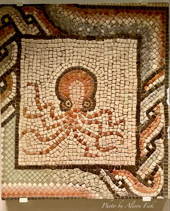 My photo shows a mosaic fragment from the floor of a Roman villa in Spain, dated AD 100-200s. It depicts a stylised octopus using red, yellow, white, and black limestone tesserae, against an off-white background. The front-facing octopus appears almost cartoon-like, with a large red and white head and body outlined in black. Its yellow circular eyes are outlined in black with a central black dot which stare out at the viewer. Below the head and body are eight writhing arms.

This octopus and other fragmentary animal mosaics were discovered in the 19th century at the Hermitage of Santa Colomba, Villaquejida, León, which had been built on the site of a Roman villa.