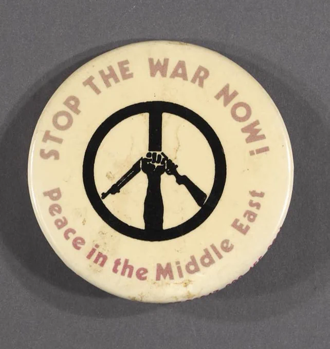 text surrounding peace sign formed by arm with fist clutching a broken rifle on a circular button 