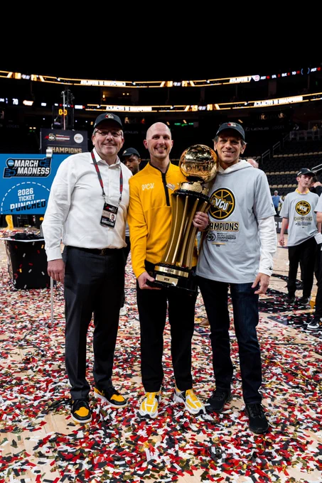Coach Martelli poses in the center with a large trophy, with Ed McLaughlin and President Rao on either side of him. They are in a basketball stadium and the floor is full of confetti.