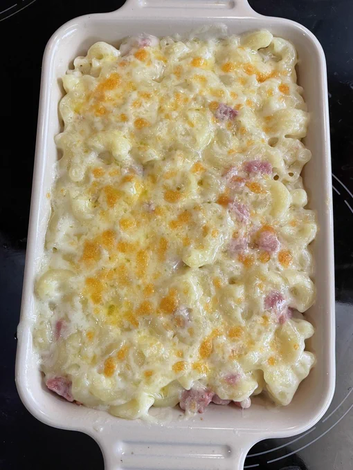 An oven proof dish on an black hob. The dish contains macaroni cheese with small chunks of pancetta with lightly browned cheese on top.