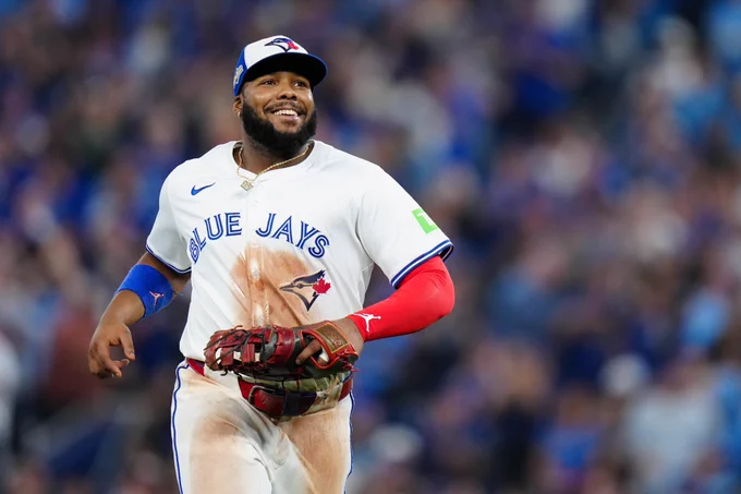 Vladimir Guerrero Jr. smiles while running on the field. He wears a white Toronto Blue Jays uniform with blue lettering and a blue and white Toronto Blue Jays cap.
