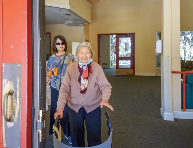 Older woman smiling and walking towards the camera using a walker. A younger woman is behind her holding bags