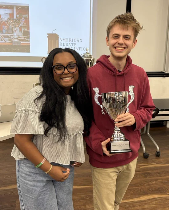 Image of Michigan State University Debate teammates Zaria Jarman and Glenn Scully standing and smiling at the American Debate Association National Championship. Jarman is leaning toward Scully, who is holding the shiny Front Royal Cup trophy.