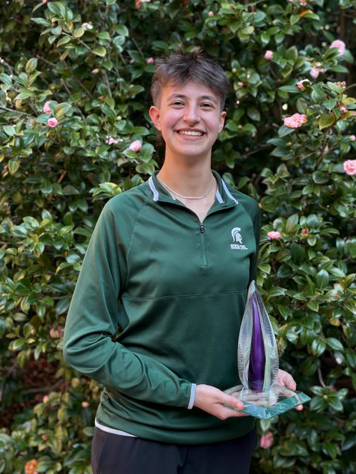 Image of Michigan State University student and debater Joanna Gusis standing and smiling while holding the glass Julia Burke Award. Gusis is wearing a dark green MSU quarter-zip, and flowering greenery is in the background.