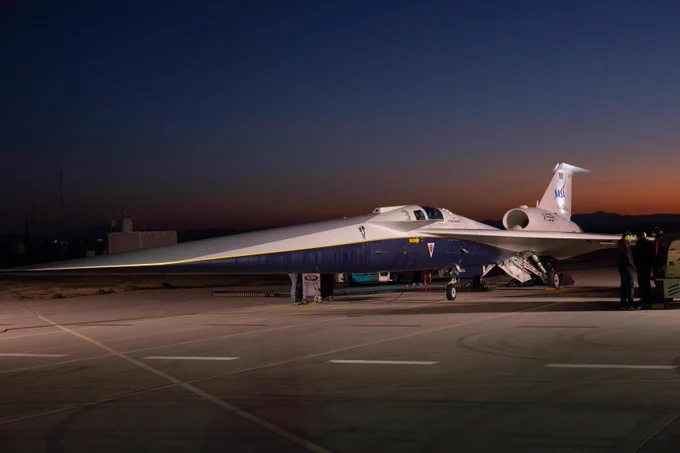 NASA's X-59 quiet supersonic aircraft, sporting a very long, aerodynamic nose, sits on the tarmac against a twilit sky. Three black-clad technicians stand near its left wing. Credit: NASA