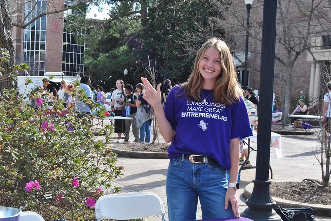 A person stands outdoors, smiling, and gesturing axe em with their hand. They are wearing a purple "Lumberjacks Make Great Entrepreneurs" T-shirt and jeans. In the background, people are gathered near a brick building, with trees and flowers around.