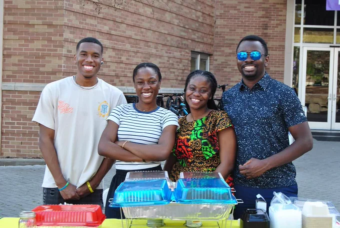 Four people are smiling and standing behind a table with trays of food outdoors. They are in front of a brick building.