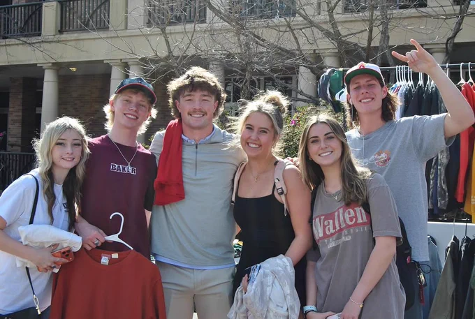 A group of six people smiling and posing together outdoors in front of a building. Some are holding clothing items on hangers. 