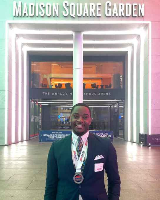 A person stands smiling in front of Madison Square Garden, wearing a suit and a medal around their neck. The entrance is illuminated with bright lights.