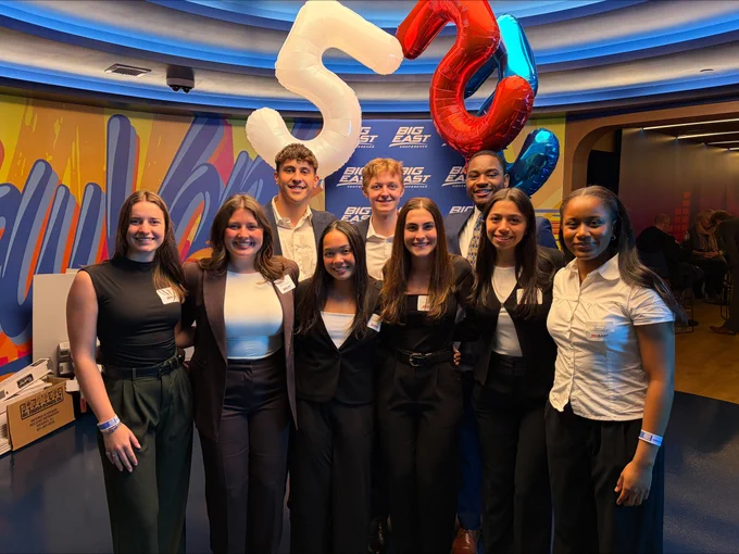 A group of eight individuals stands together, smiling, in a room with a colorful background and "Big Ten" logo. Two are holding balloon numbers "5" and "2." They are dressed in formal and semi-formal attire.