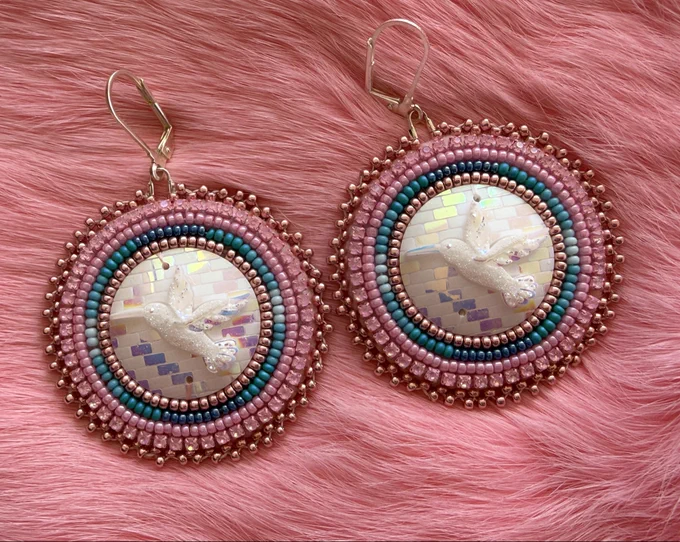 Pink and turquoise beaded flat stitched earrings with white resin cabochon centers with hummingbird design. Laying on pink rabbit fur.