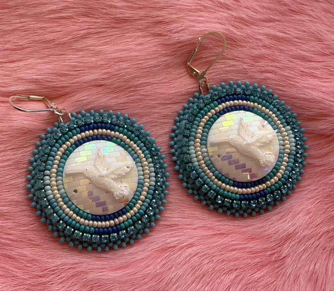 turquoise beaded flat stitched earrings with white resin cabochon centers with hummingbird design. Laying on pink rabbit fur.