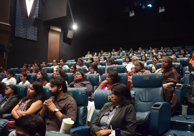 A cinema hall filled with a diverse audience seated in blue recliner chairs, attentively watching the screening of Guthlee Laddoo.
