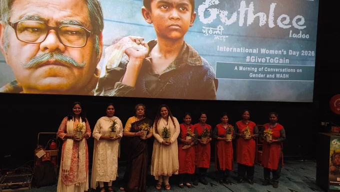 A group of women stand on stage in a cinema hall, holding small potted plants, in front of a large screen displaying the “Guthlee Ladoo” film poster for International Women’s Day 2026.
