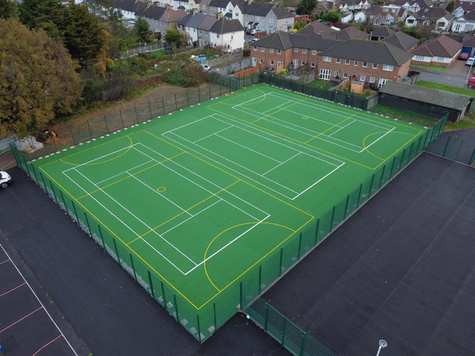 Aerial view of a fenced multi-sport court with green surface and white and yellow markings, surrounded by residential houses and trees.