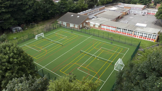 Aerial view of a fenced multi-sport court with soccer goals and various court markings beside a school building.