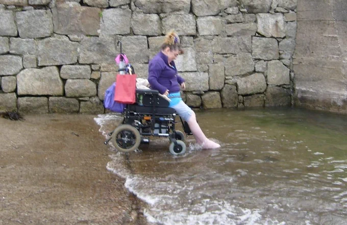 Penny, a power wheelchair user, is on a stone ramp, paddling her feet in the Irish Sea, in a little town south of Dublin. She wears a purple cardigan and blue leggings rolled up to the knees. Her hair is in a bun, brown and blonde. It’s a side view, and she’s smiling. Behind her is a wall made of large grey stone blocks.