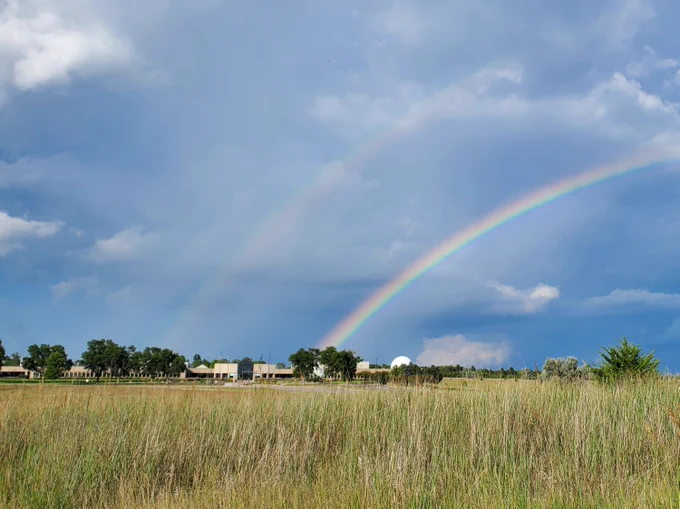 Bright double rainbow arches over a rural landscape with tall grass and scattered trees under a cloudy sky.