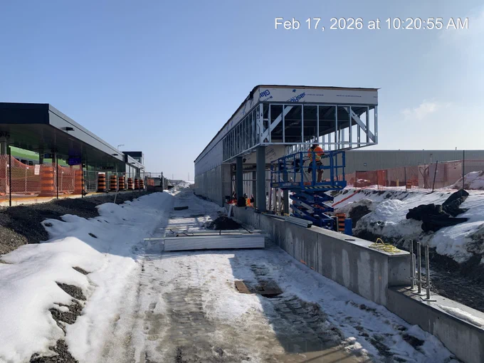 Chantier de construction d’une station de transport en commun où l’on aperçoit des ouvriers sur une nacelle élévatrice bleue. La structure du bâtiment est partiellement achevée. Il y a de la neige au sol, et des barrières de sécurité recouvertes d’un filet orange bordent les côtés. Le ciel est clair et dégagé.