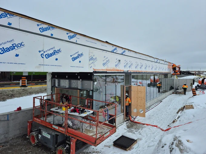Des ouvriers du bâtiment installent de grandes baies vitrées sur un immeuble. Le bâtiment est recouvert d’un matériau de la marque GlasRoc. La neige recouvre le sol autour du chantier. Une nacelle élévatrice rouge est utilisée pour accéder aux parties les plus hautes de la structure. Des cônes de signalisation et des panneaux orange sont disposés à proximité.
