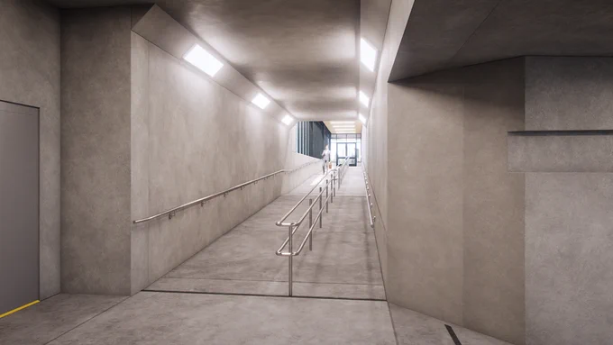 Un couloir intérieur moderne et bien éclairé, en béton, doté d’une rampe en pente douce avec des rampes d’accès de chaque côté. Les murs et le plafond sont en béton lisse. Des lampes fluorescentes éclairent le passage, qui mène à une porte vitrée au fond. Des lignes au sol indiquent le chemin accessible.