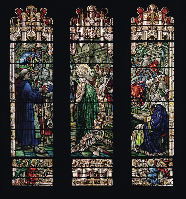 A stained glass window depicting three panels showing religious scenes. Figures in robes are engaged in various activities, such as reading and holding religious symbols. The design includes intricate details and vibrant colors, with a prominent use of blues, greens, and reds.