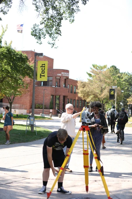 A group of students and an instructor use a surveying instrument on a campus sidewalk near a building with a Michigan Tech banner. Pedestrians and cyclists pass by as measurements are taken.