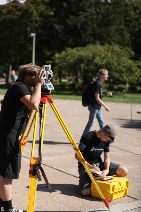 A student looks through a surveying instrument mounted on a yellow tripod while another records notes on a notebook nearby. Other students walk across a sunny campus walkway in the background.