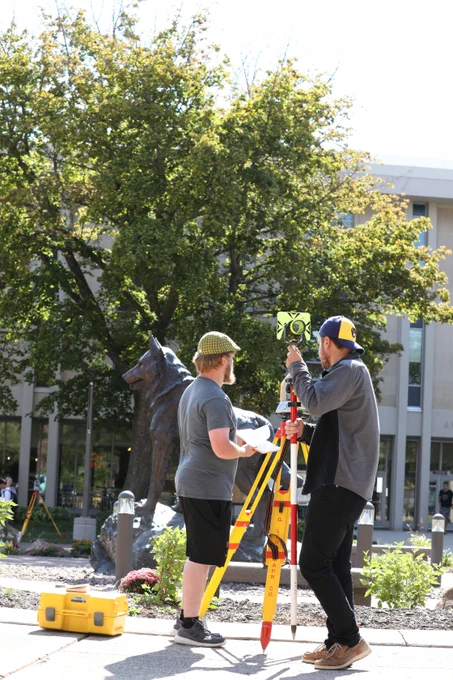Two students set up and use a surveying instrument on a tripod near a campus statue of a husky. One adjusts the equipment while the other holds notes, with trees and buildings in the background.